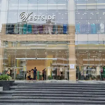 Create a realistic daytime photograph-style image of a modern Indian high-street shopping market in Sector 75, Noida. Show a clean two-storey retail storefront with large glass windows, clothing displays visible inside, a wide footpath, a few parked scooters, and a mall building visible across the road. The storefront sign should read exactly “WESTSIDE” in simple white lettering on a dark blue signboard. Use natural daylight, realistic Indian urban surroundings, and a wide-angle eye-level view. Do not include people’s faces, brand logos other than the WESTSIDE sign, or text other than “WESTSIDE”.